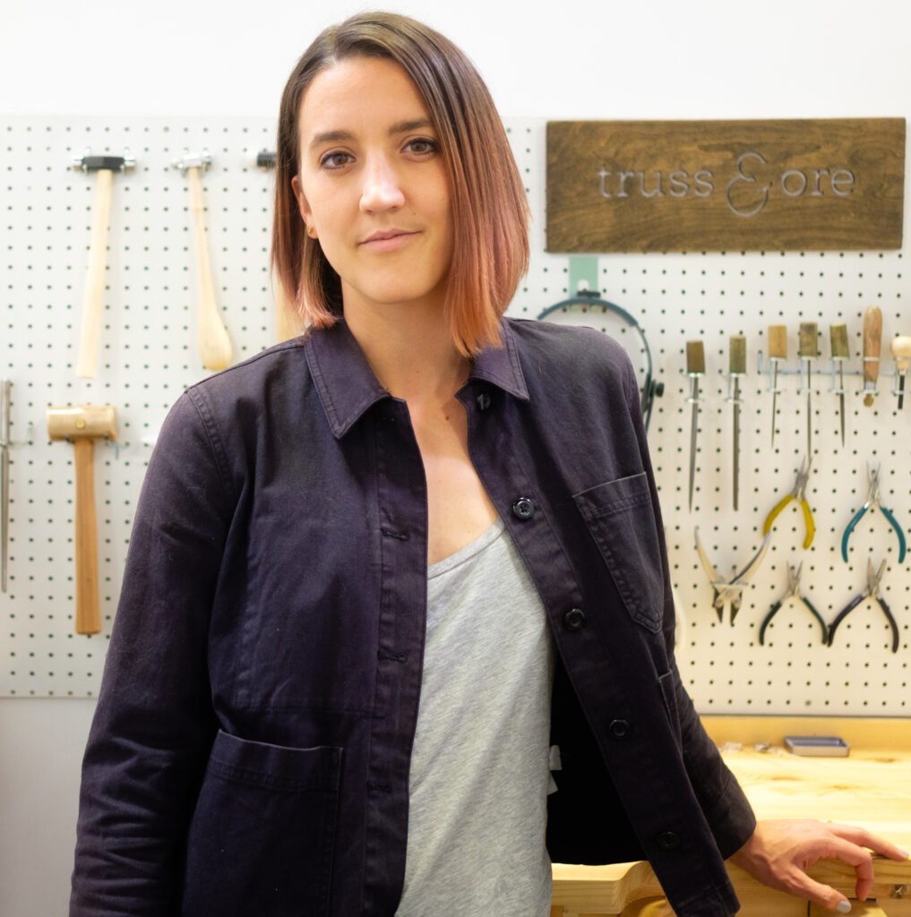 Portrait of Ana Brazaityte, standing at a jewelry bench in front of a wall of jewelry tools