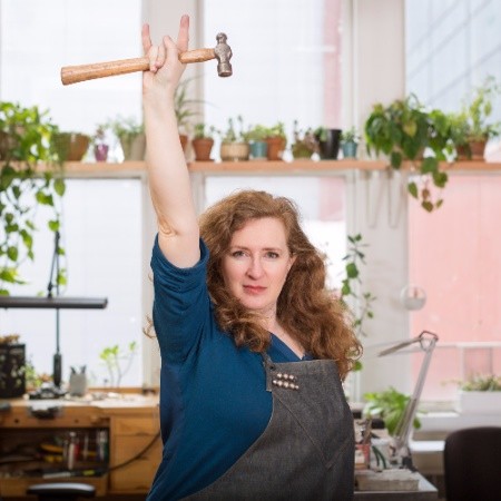 Portrait of Sharon Zimmerman in her studio, holding a hammer in her raised hand while giving the rock and roll hand sign